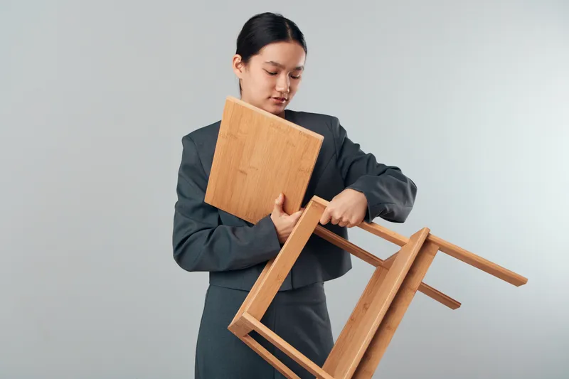 Businesswoman in suit holding wooden chair and tabletop indoors with neutral background for office furniture, workspace setup, and modern interior design concept.