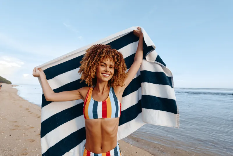 Beach girl with curly hair waves a striped flag blue and white by the ocean on a sunny seaside shoreline, joyful carefree summer holiday mood, standing near waterline with summer breeze