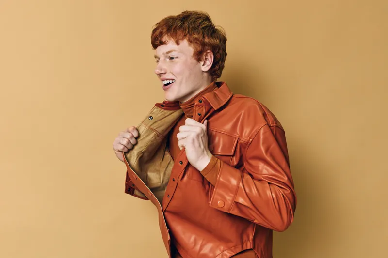 Young man wearing orange leather jacket posing against beige background, smiling and showing confident expression with red hair and casual style in studio.
