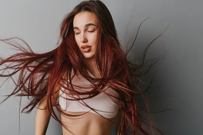 Woman with long red hair and closed eyes in motion wearing white crop top on gray background. Female model with flowing hair and natural expression in studio for beauty and fashion.