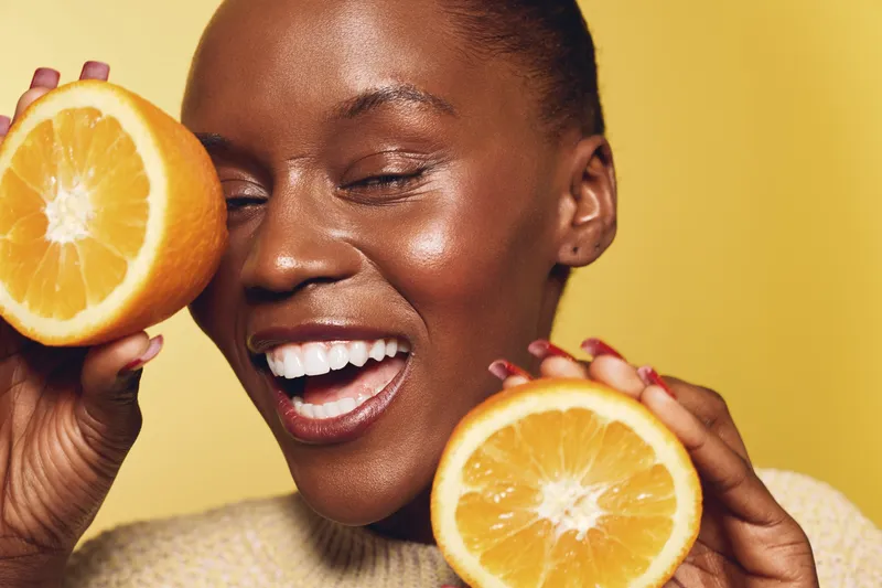 Happy African woman with dark skin holding two orange halves near face, smiling with closed eyes. Yellow background and natural lighting create fresh and joyful mood for healthy lifestyle concept.