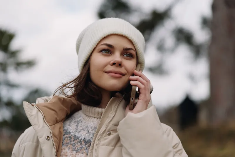 Woman outdoors wearing a warm beige jacket and knit beanie, talking on a smartphone amid a chilly park setting, soft light and distant trees, candid portrait capturing everyday communication and