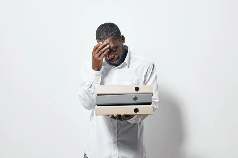 African man with dark skin holding folders and touching forehead in stress or headache. Businessman in white shirt with documents for office work and tired expression on face.