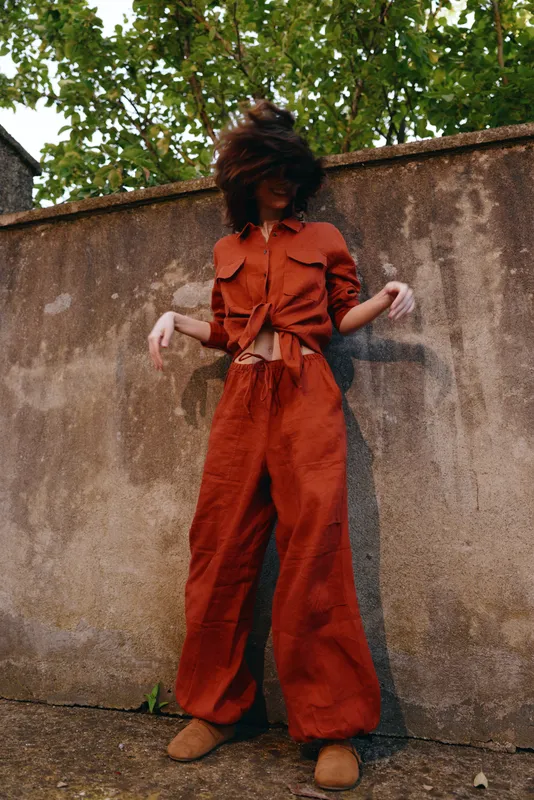Vibrant woman in rust overalls outdoors, joyful movement, windy hair, sunlit courtyard, casual lifestyle portrait with relaxed pose and natural earthy tones