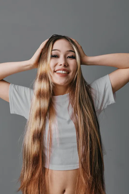 Young woman with long straight hair smiling and posing with hands on head studio portrait against gray background. Casual appearance wearing white cropped t shirt and relaxed expression