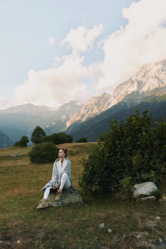 A woman sits on a rock in an open meadow with distant mountains, calm and reflective. A lifestyle portrait showing outdoor serenity and nature inspired mood.