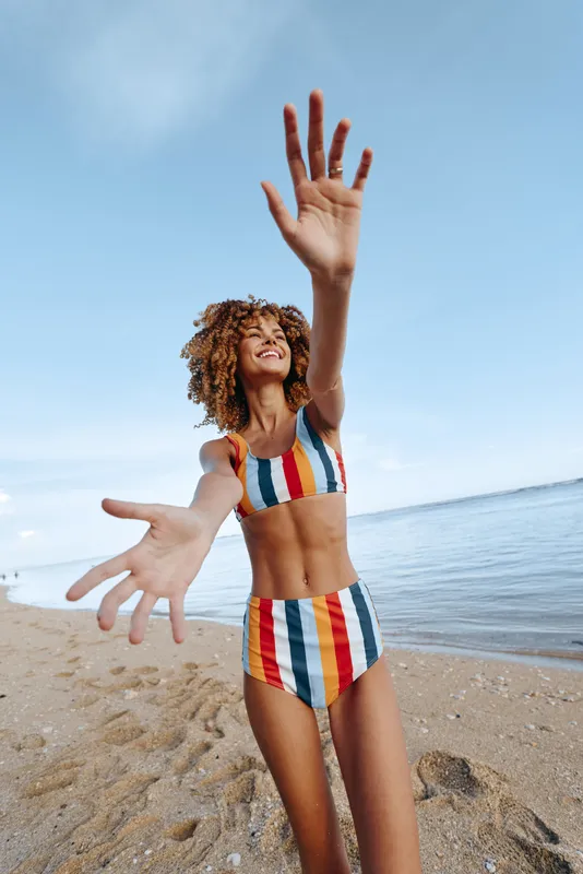 Beach, girl, swimsuit, summer, ocean, sky, joy, curly hair, sunlight, vacation, child plays on sandy shoreline, raised arms and open hands toward ocean, carefree summer fun