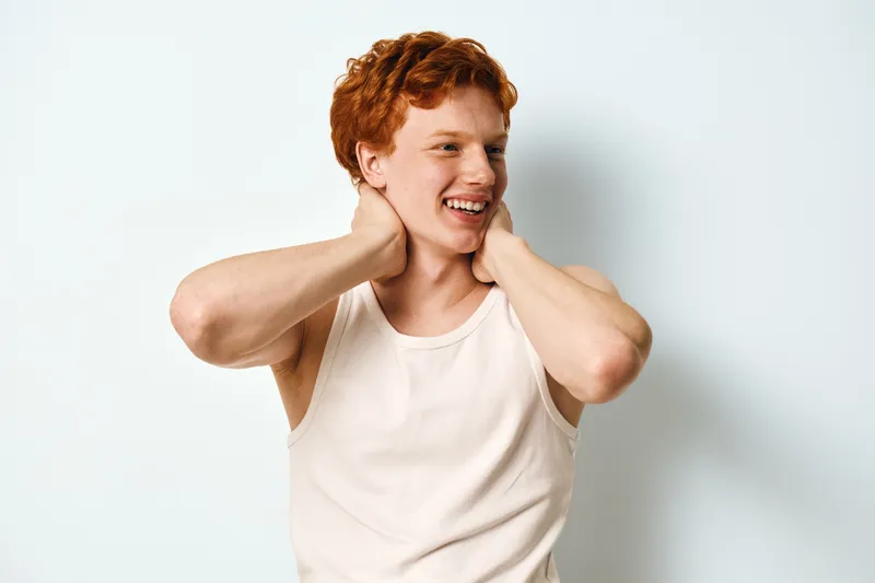 Young man with red hair and freckles in white tank top smiling and touching neck in casual studio portrait with white background and natural expression.