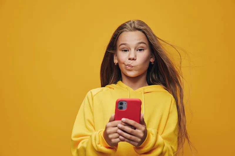 Girl with long hair wearing yellow hoodie holding red smartphone and making funny face with puffed cheeks isolated on bright yellow background for communication fun.