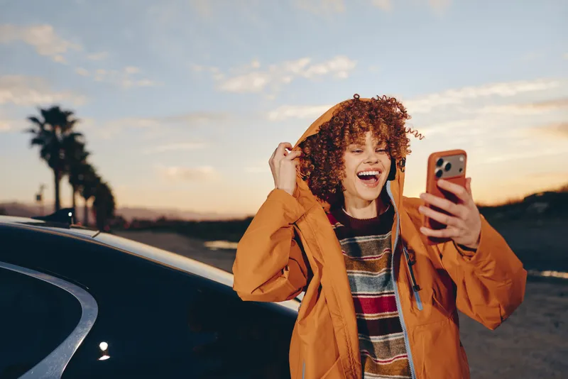 Woman lifestyle outdoors at sunset, laughing with a smartphone, wearing orange jacket, joyful face, standing by a car near roadside, warm evening sky, natural colors, bright smile