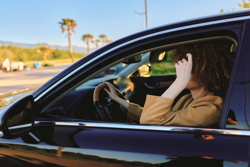 A woman sits in a car with her head resting on her hand, looking away in bright daylight. Concept for stress, burnout, and mental health in everyday life and driving.