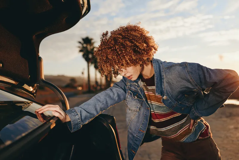 Woman lifestyle, car road sunset, denim jacket, curly hair, outdoor scene, person leaning into vehicle, holding door handle, travel mood, warm light