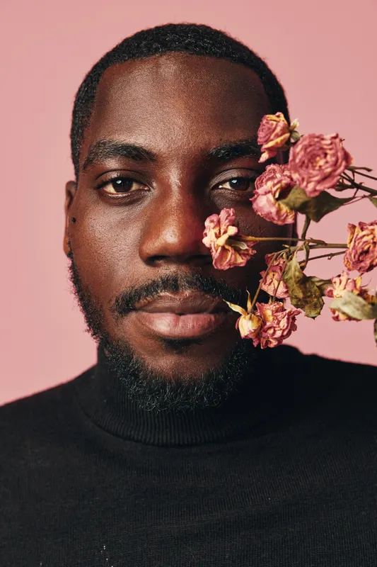 African man portrait with dried pink flowers on face in creative conceptual visual style. Closeup, black sweater and pink background for artistic expression and beauty.
