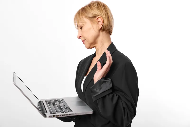 Woman in office clothes shows stress and anxiety, holding her neck with pain and strain while using a laptop during computer work, tense posture, profile view on white