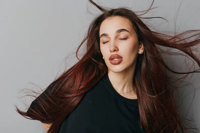 Woman with long hair and closed eyes posing with wind blowing through hair for beauty, fashion, and hairstyle concept in studio with neutral background and black shirt.