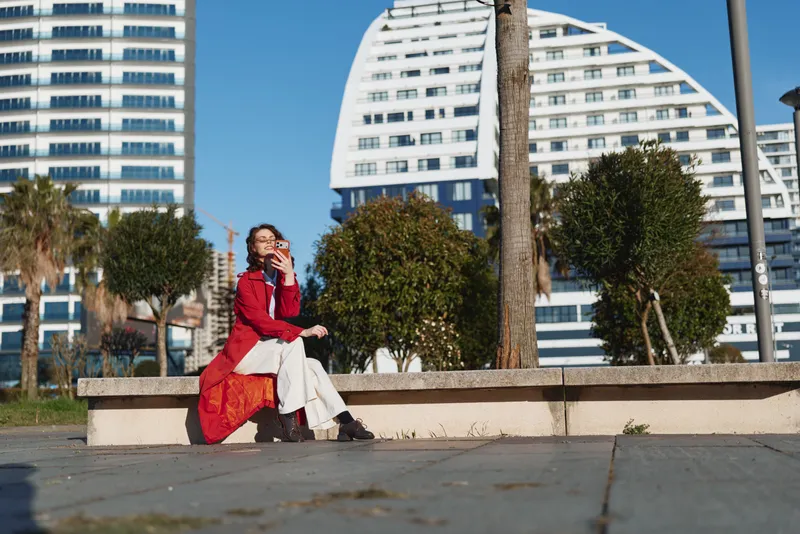 A young woman in a red coat sits on a concrete bench in a modern city plaza, taking a selfie with her phone and smiling brightly today.