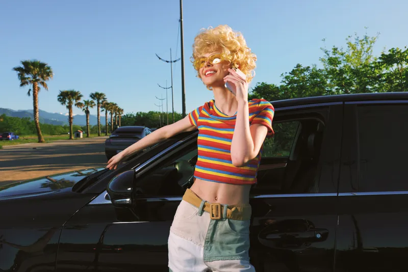 A young woman with a retro striped crop top and blonde wig leans on a dark car, smiling, in a sunny coastal street. Vintage lifestyle, bold color and carefree mood.