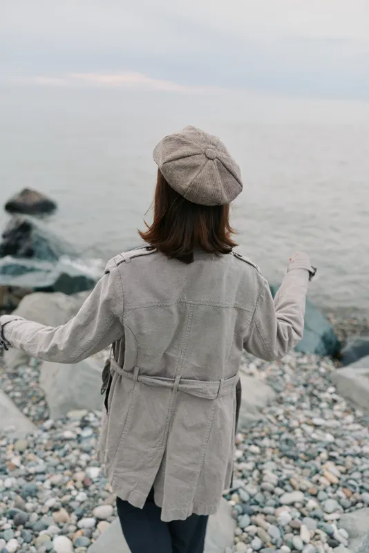 Woman in beige coat and hat stands on rocky beach facing calm sea. Overcast sky and muted tones create a peaceful and contemplative mood by the water. Concept of solitude and nature.