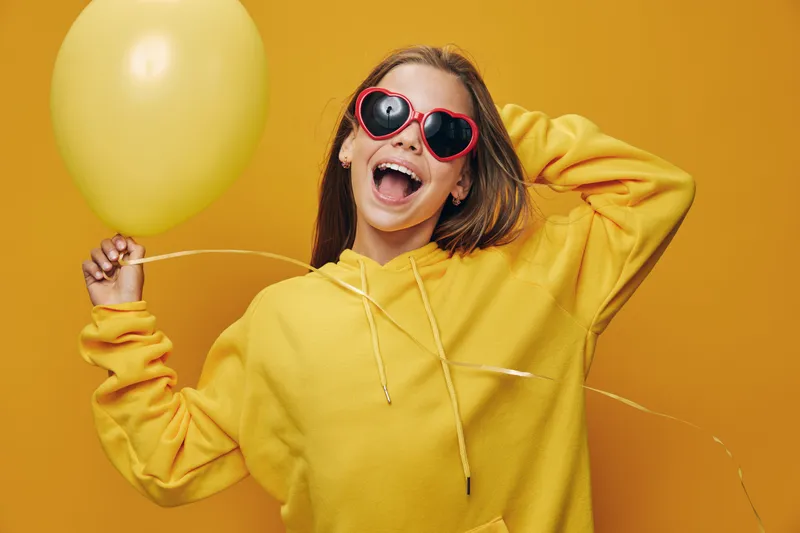 Happy woman in yellow hoodie and red sunglasses holding balloon, smiling with excitement against yellow background for celebration and fun mood.
