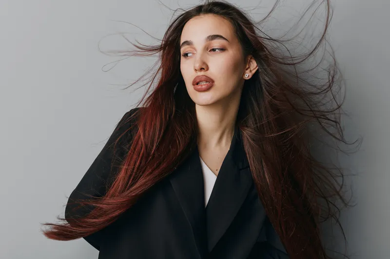 Young woman with long flowing hair wearing black coat posing against gray background. Fashion portrait with natural makeup and confident expression for beauty and style concept.