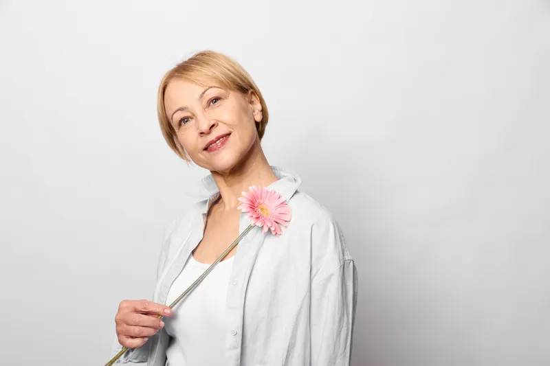 Woman portrait in studio, smiling with relaxed expression, blonde hair, pink flower detail on white jacket, soft light, clean white background, casual fashion mood