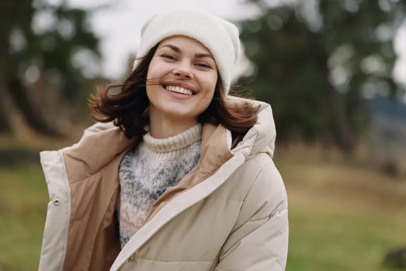 Woman smiling in an oversized beige coat and knit sweater outdoors in a park during cool weather, showcasing casual fashion and a cheerful mood for a portrait.