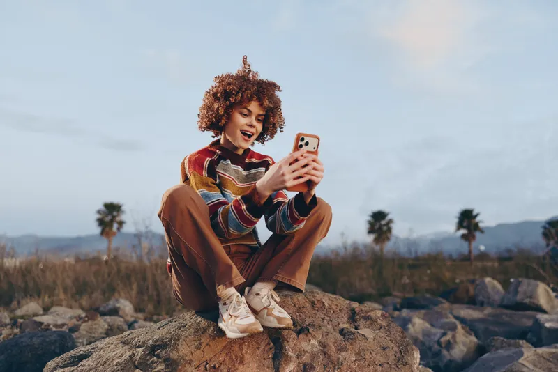 Woman lifestyle, curly hair, selfie on smartphone outdoors, sitting on a rock in nature, smiling, soft golden light, casual sweater, relaxed travel portrait, bright sky, peaceful mood