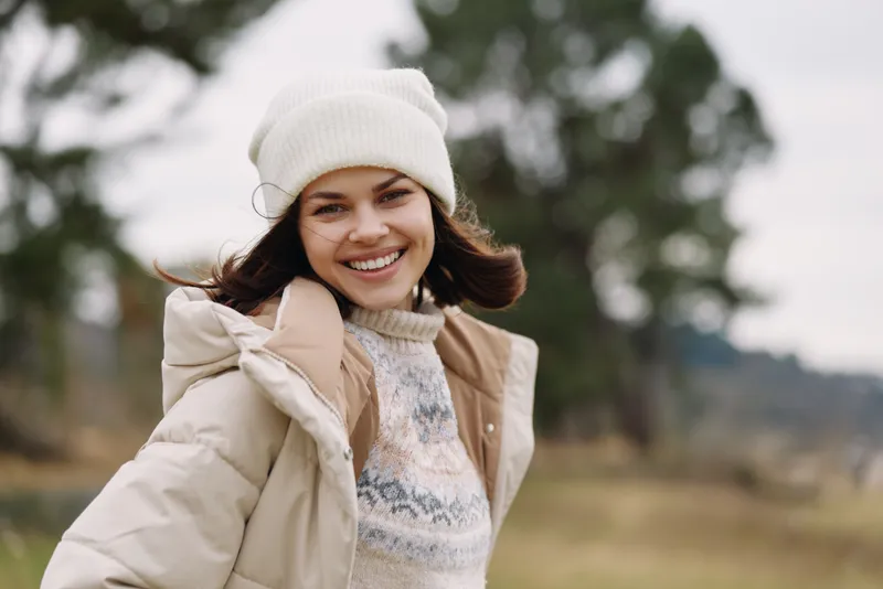 Woman in a warm beige coat and knitted sweater smiles softly outdoors on a windy winter day, wearing a white beanie, with a blurred natural background of trees and fields.