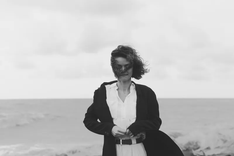 Woman stands on a windy beach wearing a dark coat over a light shirt, looking downward with a calm, thoughtful expression as waves roll in under a cloudy sky.