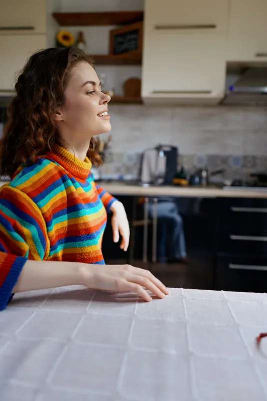 A young woman with curly hair sits at a table in a bright kitchen, wearing a colorful striped sweater and smiling softly, capturing a relaxed home moment.
