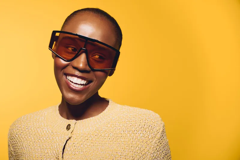 Smiling African woman with dark skin wearing large tinted sunglasses and a beige knit sweater on yellow background. Concept of fashion, style, confidence, and happiness.