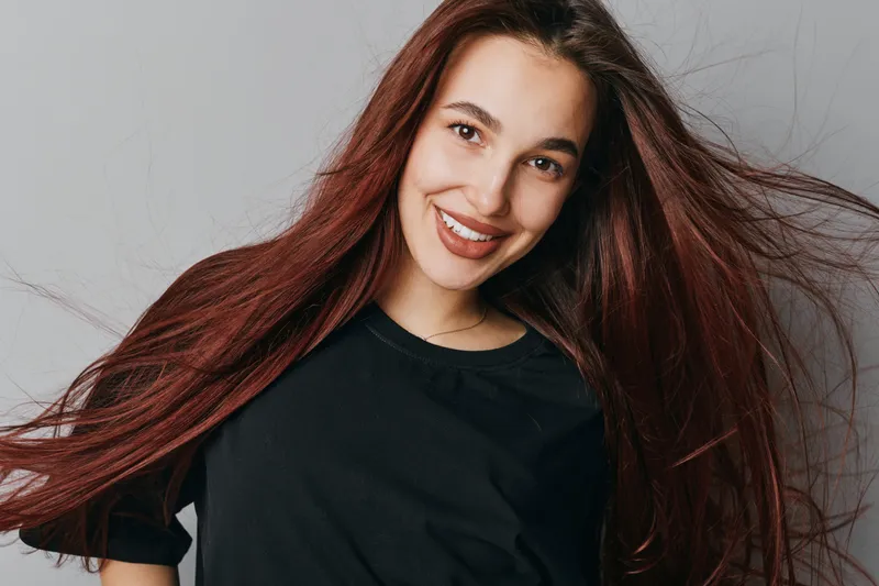 Young woman with long red hair smiling and looking at camera wearing black shirt on grey background. Portrait of happy female with natural makeup and flowing hair in studio light.