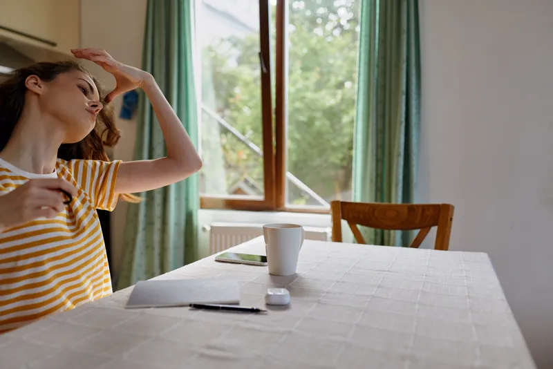 Young woman wearing a striped shirt relaxes at a sunlit kitchen table, eyes closed, enjoying warm window light and a calm, carefree moment.