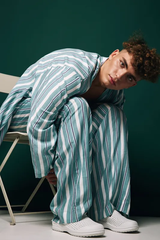 Young man in striped pajamas sitting on white chair against dark green background, casual pose with curly hair and white slip-on shoes, indoor studio portrait with relaxed mood.