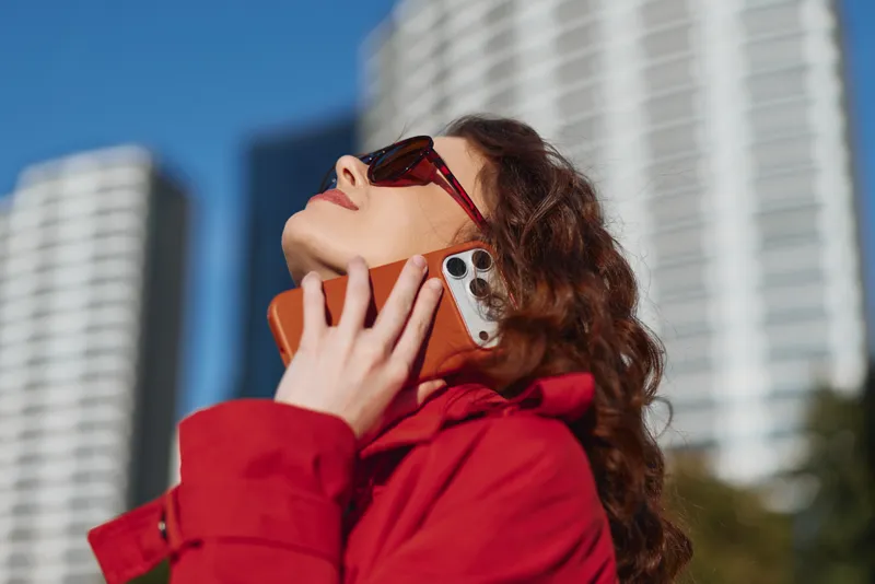 A woman in a red coat talks on a smartphone outdoors, looking up with sunglasses as high-rise buildings loom in the bright city, capturing urban communication and confidence.