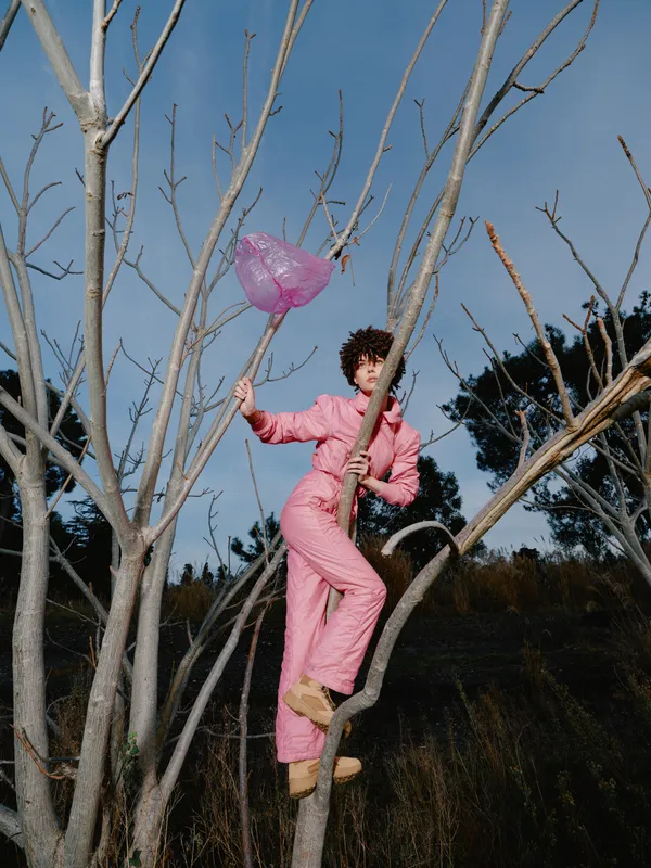Child in a pink jumpsuit climbs a bare tree with a pink balloon, outdoors in a field, capturing playful adventure and curious exploration in warm, natural light.