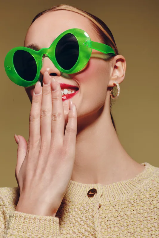 Young woman with bright green sunglasses and red lipstick covers her mouth with hand, smiling. Studio portrait with beige background and cozy knitted sweater. Concept of fashion and style.