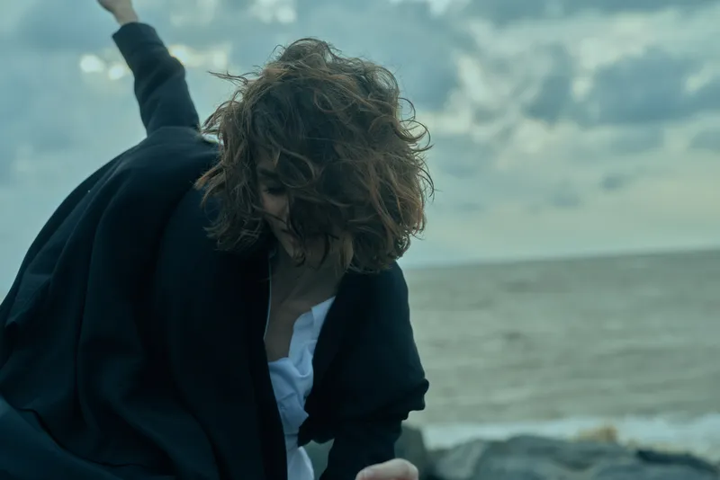 Beach scene with wind, motion, and a businesswoman in blazer and white shirt, outdoors by the sea, capturing dynamic energy and resilience against a cloudy sky