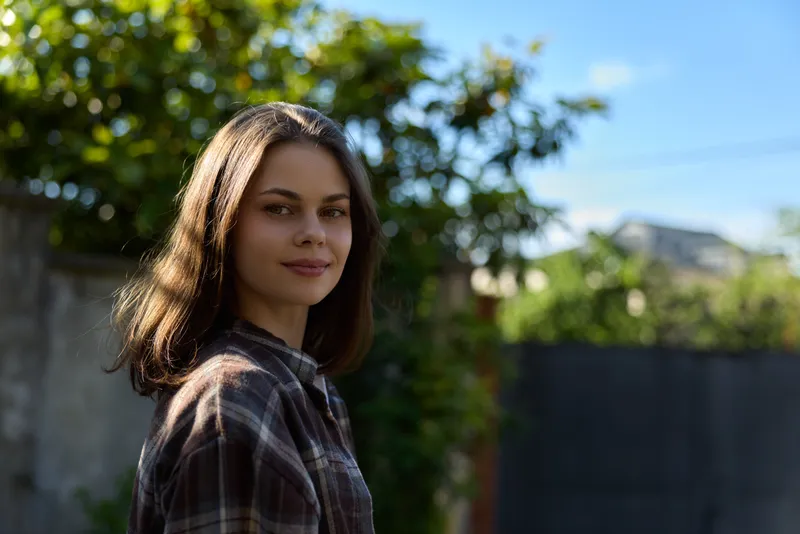 Young woman with brunette hair stands in soft sunlight, wearing a plaid shirt, turning slightly toward the camera with a gentle smile, her casual pose and warm expression convey everyday confidence