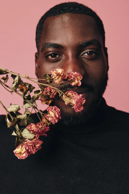 African man with dark skin and beard holding dried pink flowers near face in creative conceptual visual style on pink background. Portrait with artistic expression and floral detail.