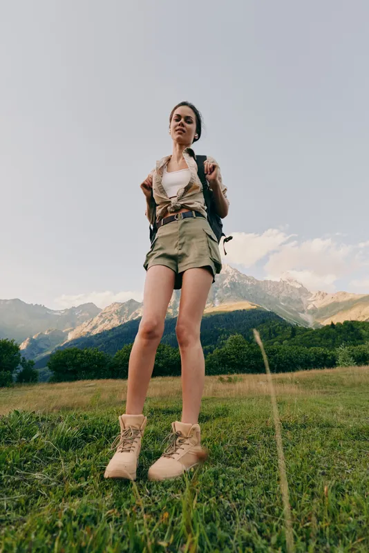 Young woman in hiking boots and shorts stands confidently on a grassy field with mountains in the background during a clear day.