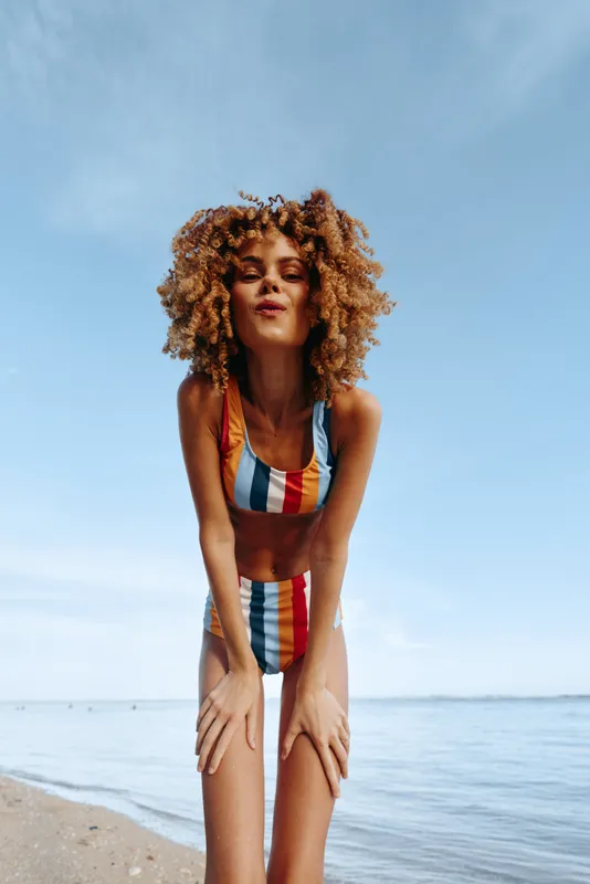 Woman with curly hair in striped swimsuit stands on sandy beach by ocean, relaxed pose under blue sky, summer outdoors lifestyle, natural beauty, serene mood