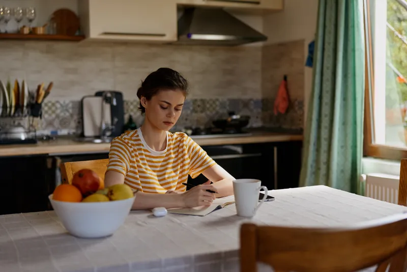 A woman in a striped shirt sits at a kitchen table using a smartphone in a cozy home setting. Natural light from a window creates a calm, everyday lifestyle moment.