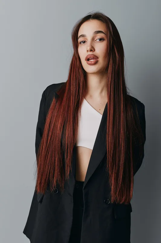 Young woman with long straight hair wearing black blazer and white top standing against gray background looking at camera with confident expression and natural makeup.