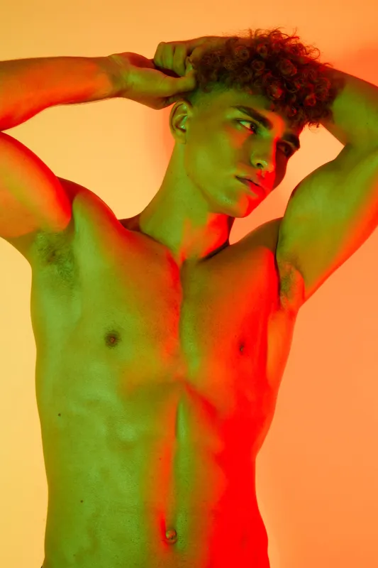 Young man with curly hair posing shirtless with arms raised, showing muscular torso and defined abs under colorful red and green lighting in studio setting.
