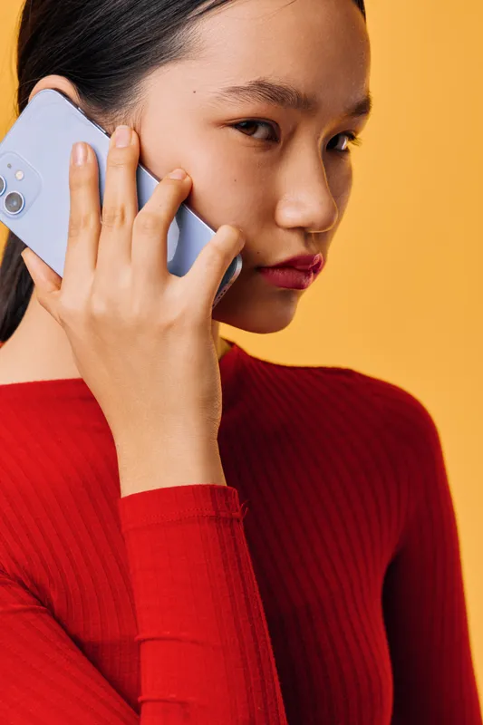 Young woman in red sweater holding smartphone to ear in closeup portrait for communication, conversation, connection and technology on yellow background with serious facial expression.