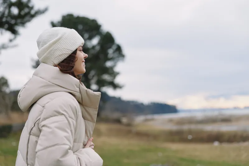 Winter woman gazing over a tranquil shoreline landscape in a puffy coat and beanie, calm breeze, soft clouds, reflective mood in an outdoor scene