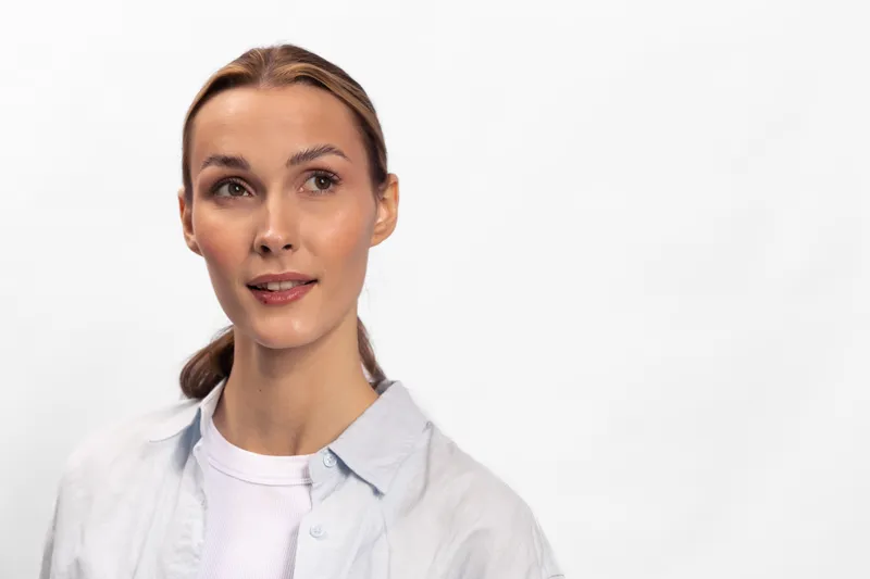 Young woman with light brown hair tied back wears white shirt and light blue button-up, looking aside on white background. Concept for casual style, beauty, and natural portrait.