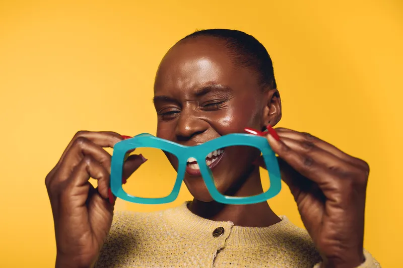 Happy dark skinned African woman smiling and holding oversized blue glasses in front of her face on yellow background. Concept of joy, fun, and playful mood with vibrant colors.