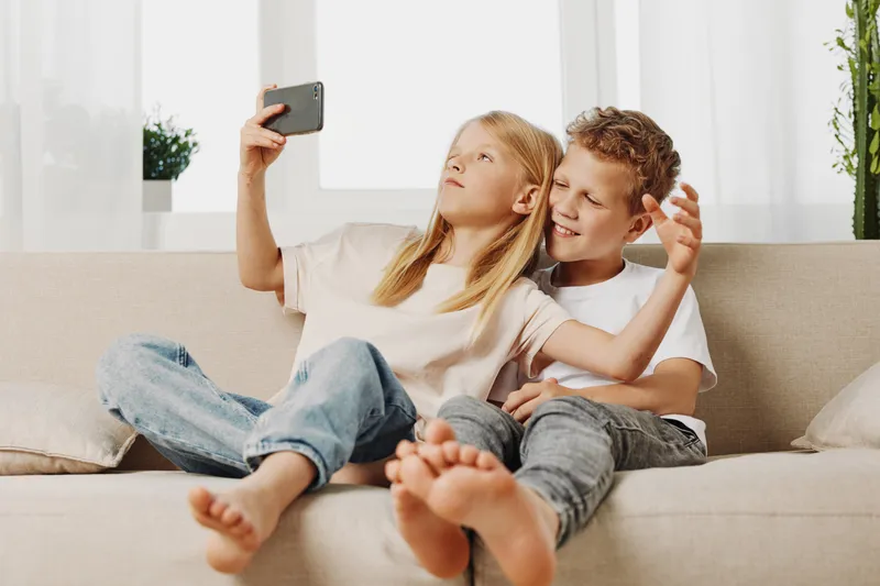 Two children sitting on a beige couch taking a selfie with a smartphone in a bright modern living room. Casual home moment with relaxed mood.